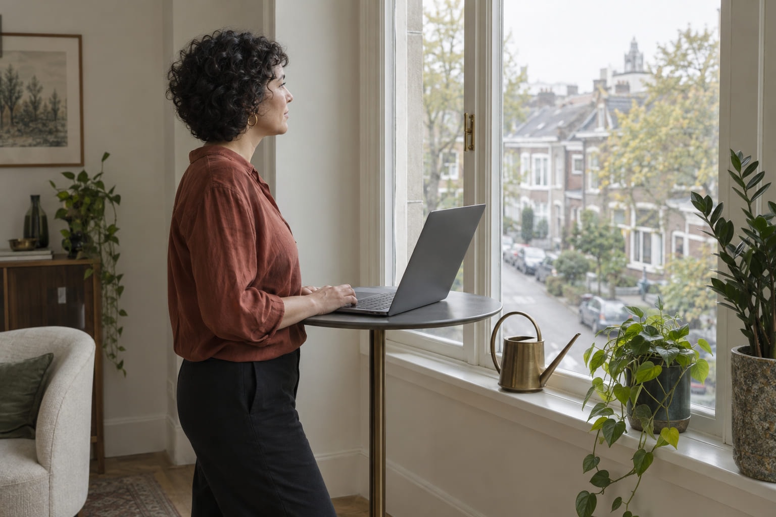 A woman standing at a window with a laptop balanced on a high table, looking out over a residential street.