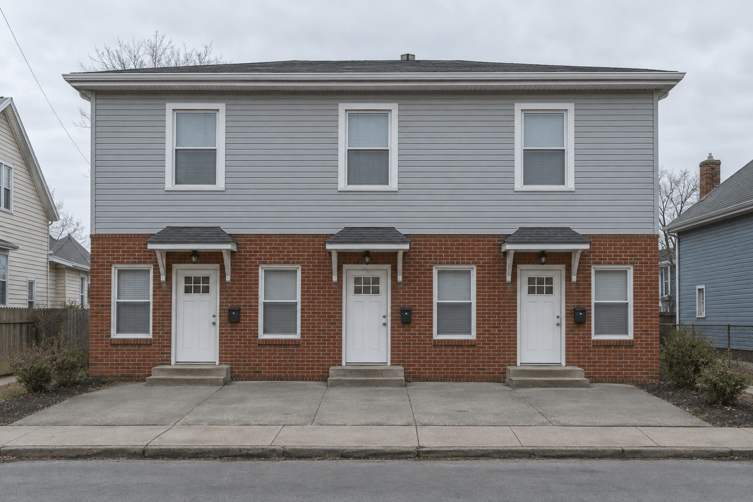 A three-unit rental building with three side-by-side entry doors, brick below and grey siding above.